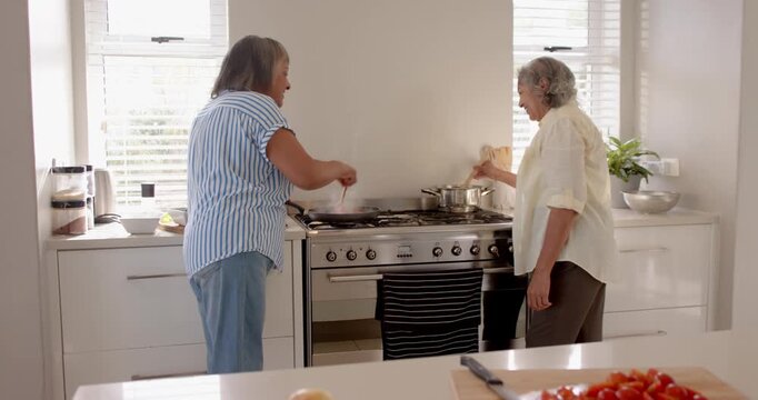Two senior female friends cooking together in modern kitchen, sharing joyful moments and laughter