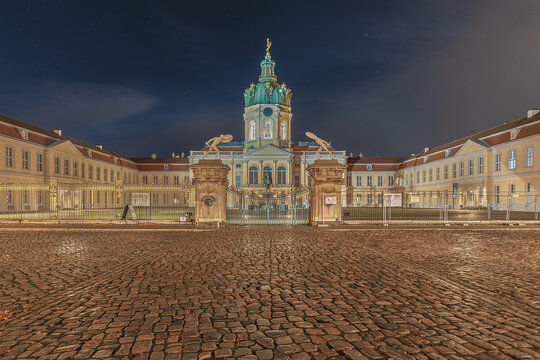 View of Charlottenburg Palace at night with its illuminated dome, ornate statues, and cobblestone courtyard in Berlin, Berlin, Germany.