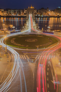 View of the illuminated Szechenyi Chain Bridge at night with vibrant light trails from traffic circling the roundabout in the foreground in Budapest, Hungary.