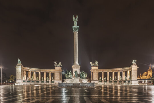 View of Heroes' Square in Budapest, Hungary, featuring the Millennium Monument with its central column and statues under the night sky, reflected on the wet pavement.
