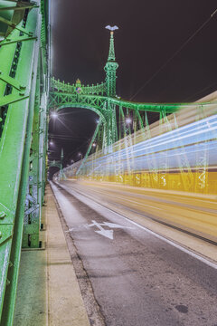 View of the Liberty Bridge in Budapest at night, featuring its iconic green iron structure, illuminated by streetlights and dynamic light trails from a passing tram, Budapest, Hungary.