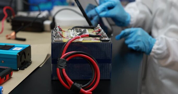 Technician in blue gloves tests a large rechargeable battery pack with red cables and electronic equipment on a laboratory workbench.