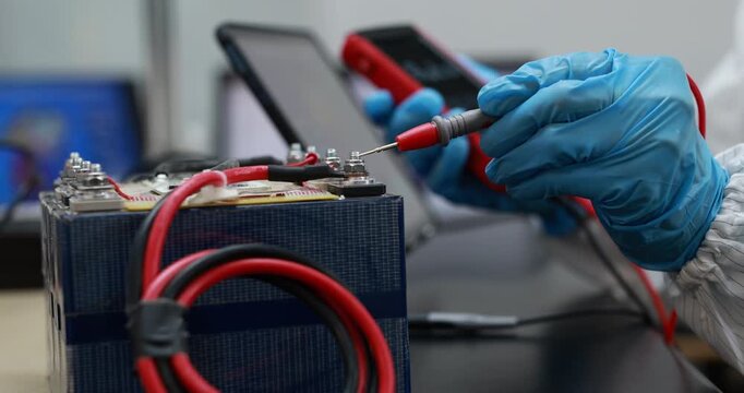 Close-up of a technician wearing blue gloves testing a battery with probes and cables in an electronics workshop.