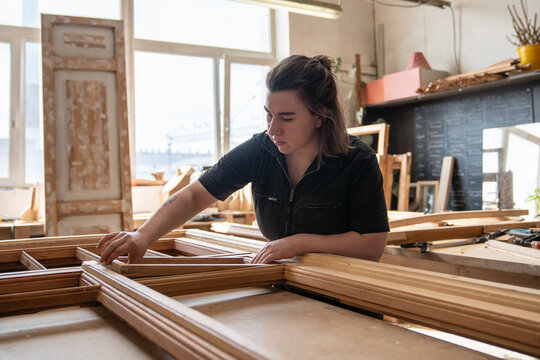 Craftswoman carefully disassembles antique wooden window frame part at workbench in workshop, woman carpenter places window section into frame structure, scrutiny and analysis of future restoration.