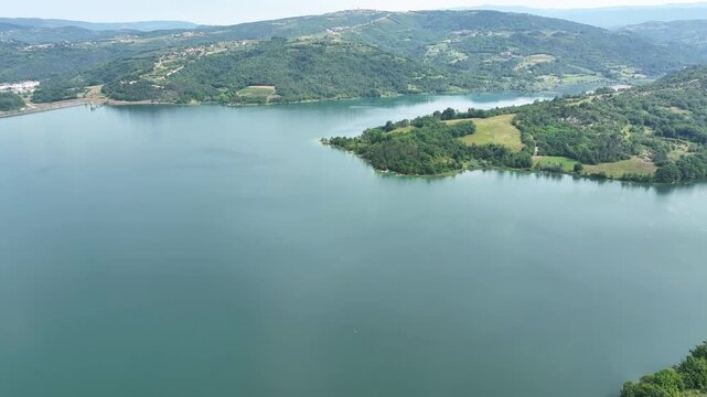 Turquoise water of Butoniga reservoir reflects white clouds in Istria region. Drone flies over artificial lake surrounded by dense green forests and rolling hills near Motovun.