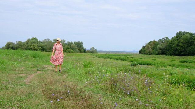 young brunette woman walks along gravel road past lush green bushes and blooming summer wildflowers. Peaceful rural landscape of Croatia offers relaxation and slow living vibes.