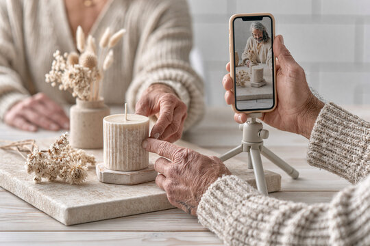 Hands arranging a handmade scented candle and dried flowers, using a smartphone snap a high-end product photo for social media.