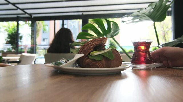 Glass of dark Turkish tea stands on wooden table next to delicious croissant sandwich. Cozy cafe interior with green plants creates relaxing atmosphere for breakfast or lunch.