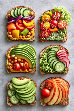 Six slices of bread topped with fresh, colorful ingredients: avocado, cucumbers, cherry tomatoes, hummus, chickpeas, bell peppers.