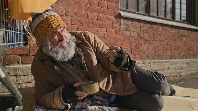 Senior Caucasian homeless man lying on sidewalk, counting coins taken from begging tin. Clip serves non-profit charity campaigns, social welfare documentaries, or poverty awareness content