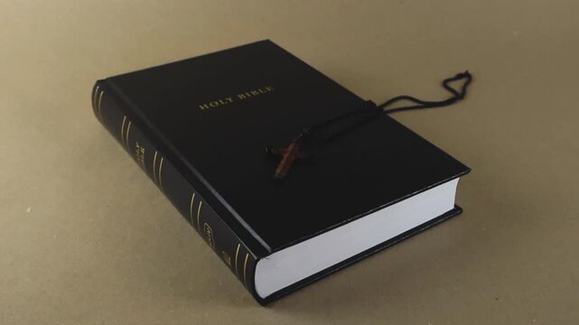 Wooden crucifix necklace falling slowly onto the black cover of the Holy Bible. The religious concept of faith, spirituality, and Christian devotion is represented by the sacred book and cross