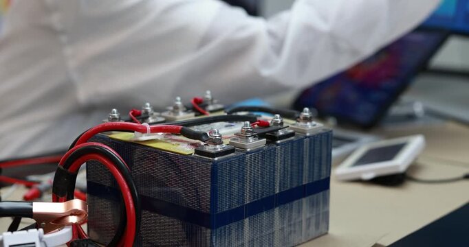Close view of a rechargeable battery pack with red and black cables on a workbench while a technician tests electrical equipment in a laboratory.