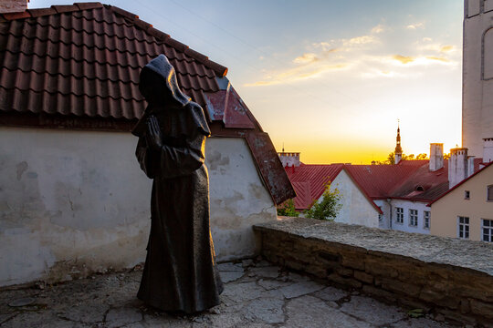 Mysterious faceless monk bronze statue at sunrise in Danish King's Garden, Tallinn, Estonia. Silhouette of medieval sculpture against a vibrant morning sky, old town roofs, and st olaf spire.