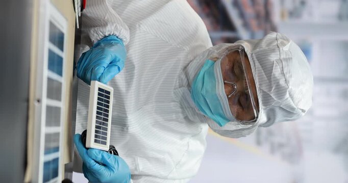 Cleanroom technician in protective suit, mask and gloves holding a small solar panel during quality inspection in a modern laboratory.