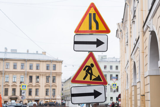 Yellow triangle road signs indicating construction work and road narrowing ahead. Group of traffic signs with directional arrow on street post against blurred historic building background.