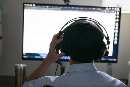 A high school student wearing headphones and doing online learning on a computer; a student working on lessons or listening exercises at home; listening to music; the student&rsquo;s back view