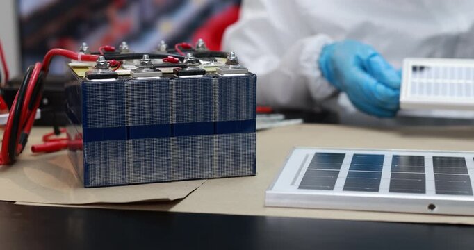 Technician in blue gloves checks a battery pack beside solar panel samples on a workbench, renewable energy testing and clean power research.