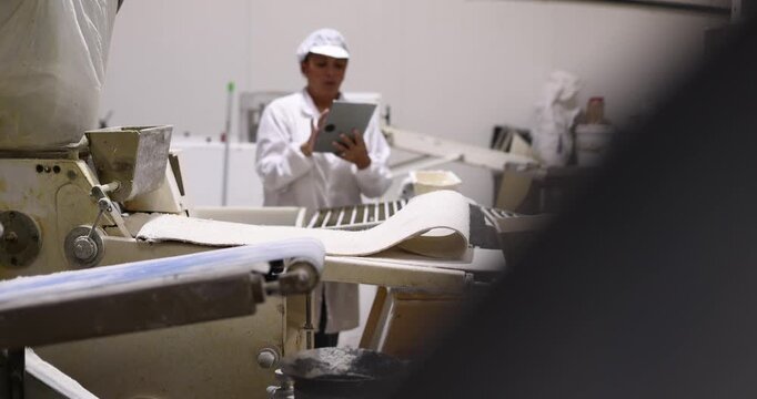 Female baker in a white uniform using a digital tablet to control the dough processing machine on a conveyor belt, ensuring quality and efficiency in a modern industrial bakery