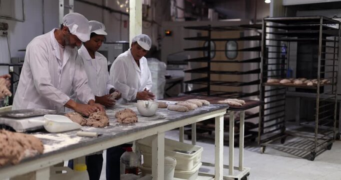 Three bakers in white uniforms and hats diligently kneading and shaping bread dough on a floured industrial table, preparing loaves for baking in a professional kitchen environment