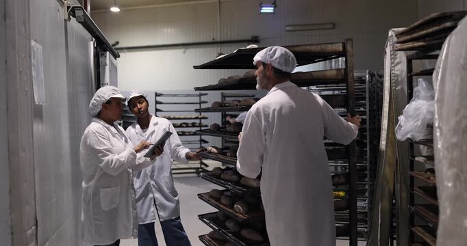Diverse team of bakery workers using a digital tablet for inventory management and quality control in a commercial kitchen, discussing production while surrounded by racks of fresh bread