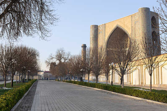 Bibi-Khanym Mosque facade and tree-lined walkway in Samarkand during early spring. The monumental iwan portal and minaret rise above a quiet brick-paved promenade bordered by green hedges.