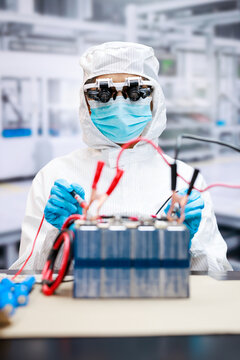 Cleanroom technician in protective gear tests a battery pack with electrical probes and wires in a modern laboratory, highlighting energy research and safety