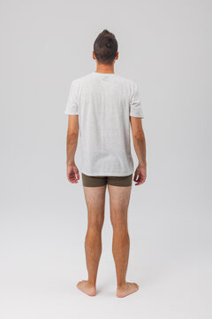 Full-length back view of a man wearing a grey t-shirt and olive-green boxer briefs. Standing barefoot on a light grey studio background. Minimalist style.