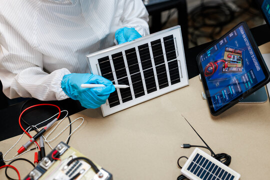 Technician in protective clothing tests a small solar panel with an electrical probe beside electronic components and a tablet in a clean laboratory.