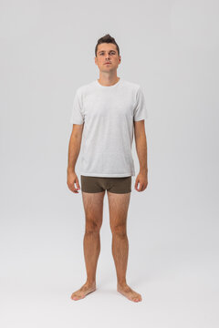 Full-length front view of a young man wearing a basic grey t-shirt and olive boxer shorts, standing barefoot against a plain studio background. Ideal for fashion and menswear mockup.
