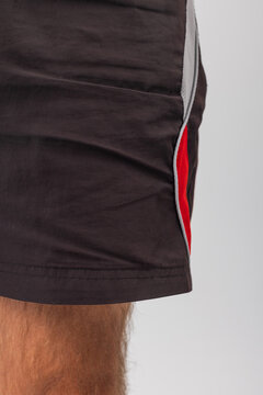 Close-up macro detail of men's black athletic shorts featuring side red and grey piping. Shows hem stitching and leg skin. Great for sportswear apparel detail and product stock photos.