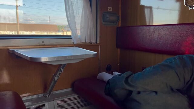 Passenger resting inside a sleeper train compartment beside a table and window. Railway travel scene illustrating long-distance transport, tourism, comfort and mobility.