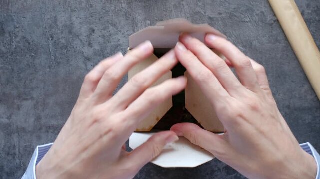 Female hands opening cardboard box with wok noodles and fresh vegetables and leaf of basil inside, ready for quick and convenient office lunch break, filmed from top down perspective on gray table