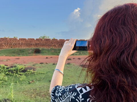 Woman with red hair taking a picture with her smartphone at the ancient Chapora Fort in Goa, India