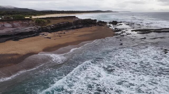 Aerial video flying over the rugged coastline at Lake Arragan, featuring sandy shoreline, rocky formations and waves rolling in from the Pacific Ocean