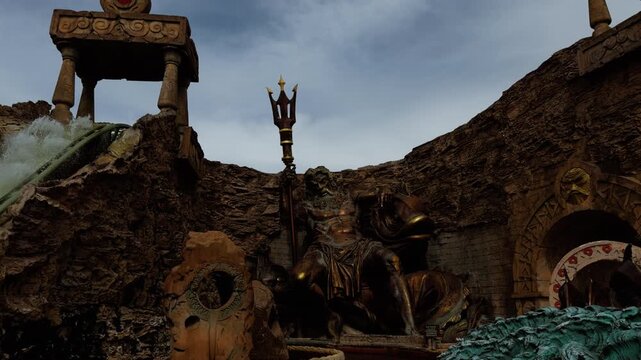 A large Neptune statue holds a trident beside the Atlantis water ride at Gardaland in Italy. The moving view shows rocky walls, columns and themed scenery.