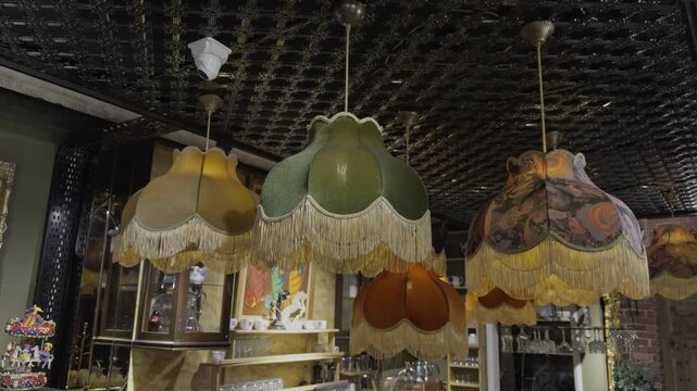 Multiple ornate scalloped fringe lampshades in green and floral patterns hanging together under a pressed tin ceiling in a vintage shop