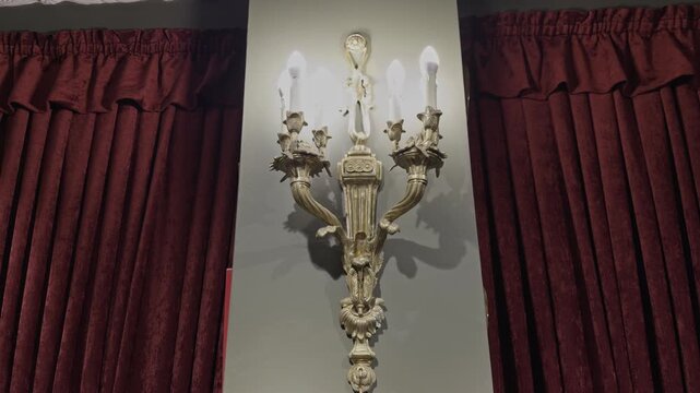 Ornate gold candelabra wall sconce with lit candle style bulbs mounted on a wall flanked by rich red velvet curtains