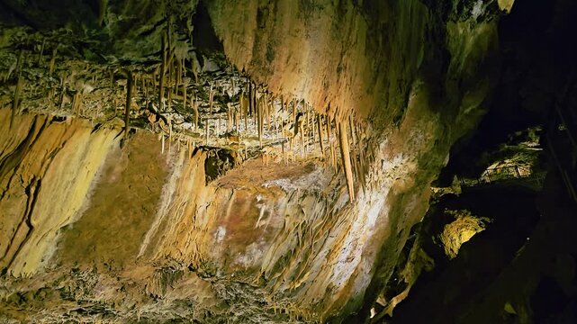 Wide interior view of a glowing underground cave with hanging stalactites, rising stalagmites, textured limestone walls, rocky pathways, and vivid blue and golden attraction lighting.
