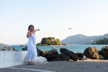 A woman in a white dress stands on a coastal pier in Kerkira, Greece, pointing toward an airplane in the sky. She reacts with an excited expression against a sea background. © dsbedarieva