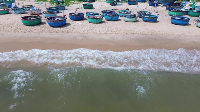 Low altitude drone perspective capturing a vibrant pattern of clustered traditional round fishing vessels and basket boats resting on the beach of Mui Ne, Vietnam.
