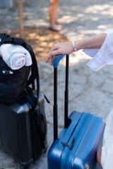 A tourist stands with multiple pieces of travel luggage on a stone pathway in Kerkira, Greece. Her hand rests on the extended handle of a blue rolling suitcase. © dsbedarieva