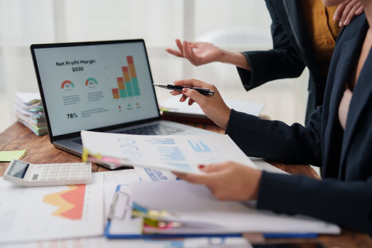 Business professionals collaborating at a desk, reviewing charts and financial reports on a laptop and printouts while discussing data-driven strategy, planning and performance
