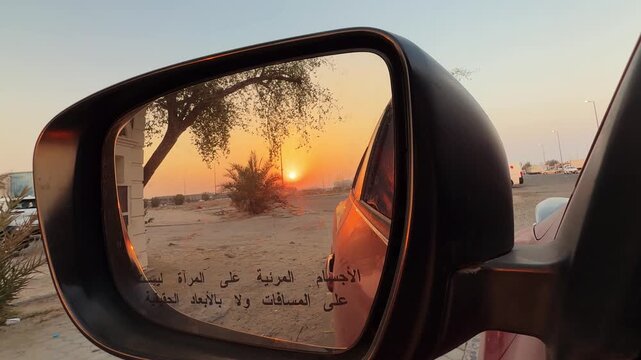 Close-up POV of a car side mirror reflecting a warm golden sunset over a sandy landscape with palm trees. Arabic warning text is visible on the glass during this peaceful evening road trip.