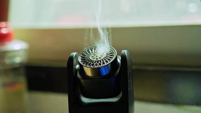 Close-up shot of a small black electronic aroma diffuser emitting a thin trail of white vapor. The device sits indoors against a softly blurred, bright daylight window background.