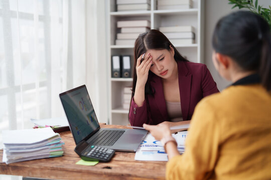 Stressed young Asian business woman feeling exhausted and having a headache, facing pressure from a female colleague in a demanding office work environment, experiencing professional burnout