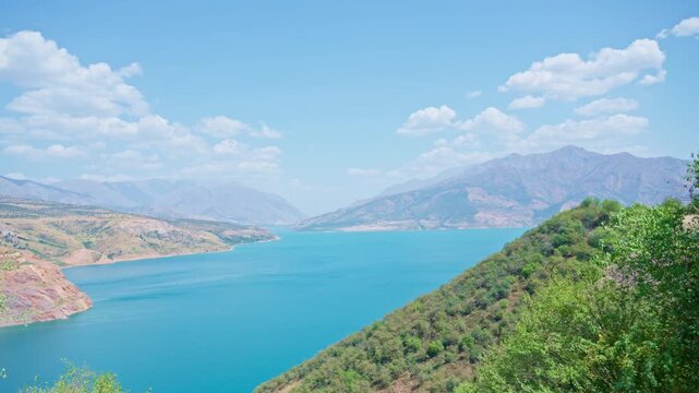 Camera moves to reveal a breathtaking panoramic view of a turquoise mountain lake surrounded by green hills under a bright blue sky with light clouds