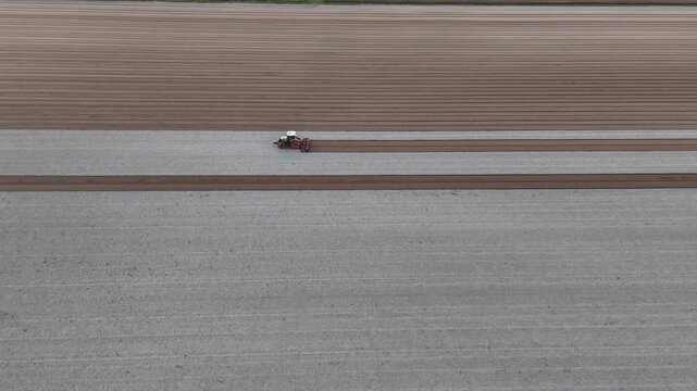 A tractor pulls an agricultural implement across freshly ploughed soil, harrowing and leveling the surface for seedbed preparation in the Po Valley of Emilia Romagna, drone tracking slide slow motion.