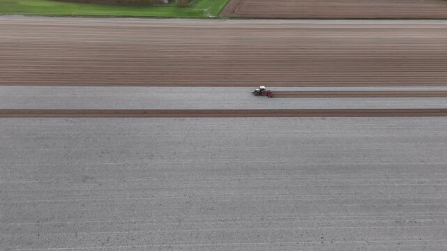 A tractor pulls a tillage implement across freshly ploughed farmland, breaking soil clods and leveling the surface for seedbed preparation in the Po Valley of Emilia Romagna, drone tracking.