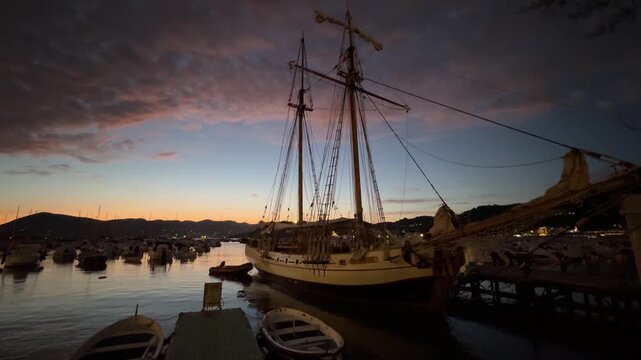 Cinematic view of historic tall ships with full sails catching the golden sunset light in the Bay of Lerici, Liguria, Italy. Silhouetted mountains and sparkling sea