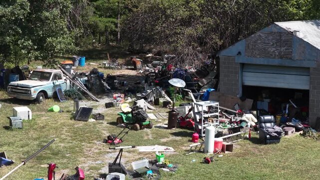 A riding mower, an armchair, a freezer and metal shelving stand among scattered belongings outside an open block garage on a rural property at Lynchburg, Virginia. Aerial view